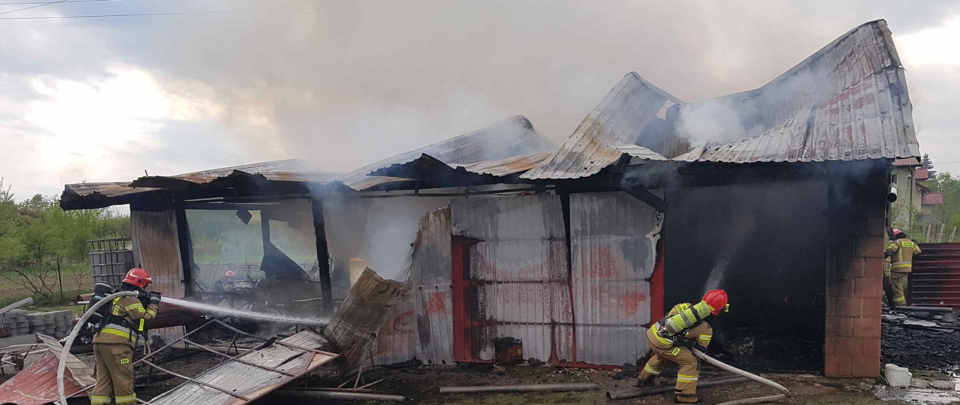Zdjęcie zrobione w dzień. Zdjęcie przedstawia palący się budynek gospodarczy. Na zdjęciu widać dwóch strażaków podających dwa prądy wody w natarciu na palący się budynek.