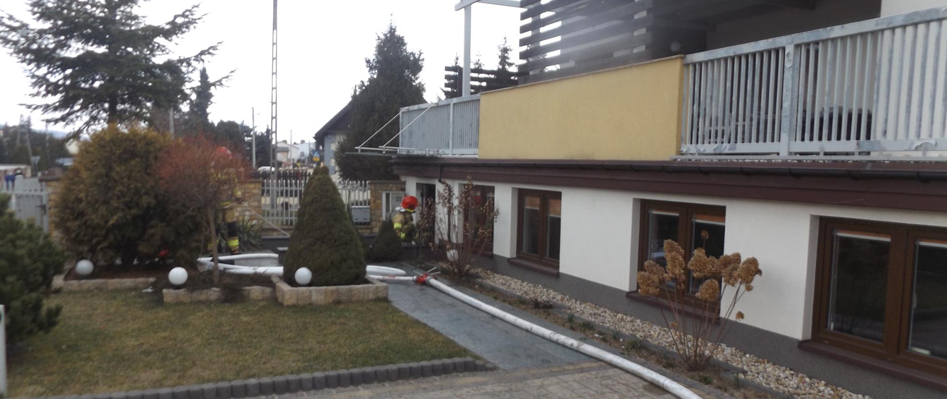 Zdjęcie przedstawia budynek w którym doszło do pożaru. Na
pierwszym planie znajduje się linia gaśnicza - za nią widać strażaka
wchodzącego do budynku. Nie widać dymu ani ognia.