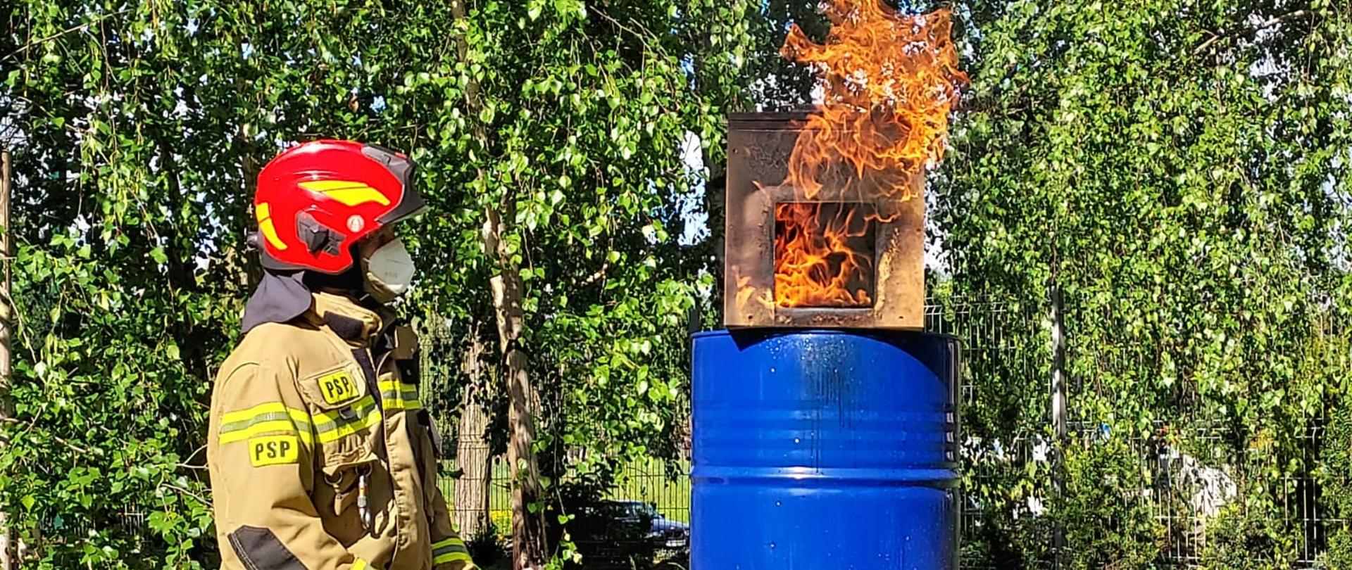 Trawnik, strażak, beczka, na beczce pali się domek z drewna