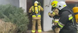Trzech strażaków ubranych w sprzęt OUO wchodzi do budynku przez drzwi wydobywa sie dym