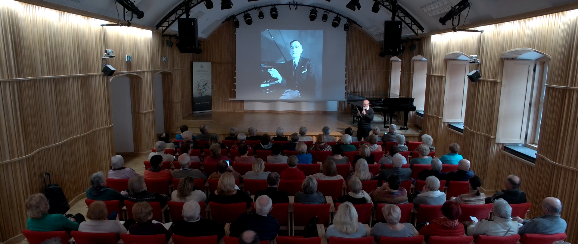 Zdjęcie. Sala koncertowa - widok od strony widowni. Na pierwszym planie widoczne od tylu rzędy słuchaczy. Przed nimi, tuż przed estradą stoi - zwrócony do publiczności - wykładowca trzymający mikrofon i kartkę. Za nim estrada, na której stoją z prawej strony dwa czarne fortepiany. Na ekranie wyświetlone jest zdjęcie kompozytora Karola Szymanowskiego. W lewym rogu stoi baner PSM Nysa.