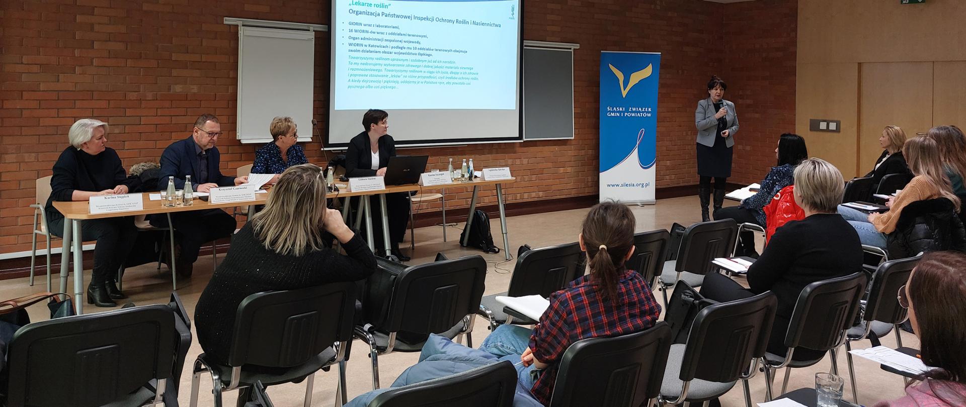 Zdjęcie przedstawia salę konferencyjną, w tle wisi rzutnik z wyświetlaną prezentacją. Na pierwszym planie czterech prelegentów siedzących przy stole, jedna osoba z mikrofonem stojąc, prowadzi dyskusję. Siedząca naprzeciwko publiczność słucha wypowiedzi.