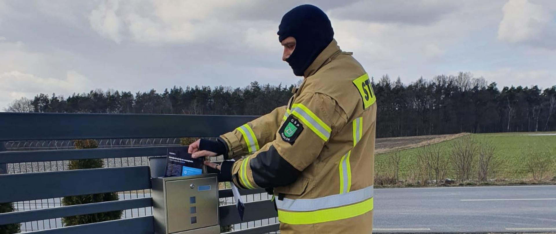 Na zdjęciu druh OSP w umundurowaniu specjalnym koloru beżowego, na głowie na czarną kominiarke. Strażak dostarcza ulotkę informacyjną o COVID-19 i szczepieniach na SARS-CoV-2 do skrzynki na listy gospodarstwa domowego. Akcja kolportażu odbywa się na terenie całego kraju.