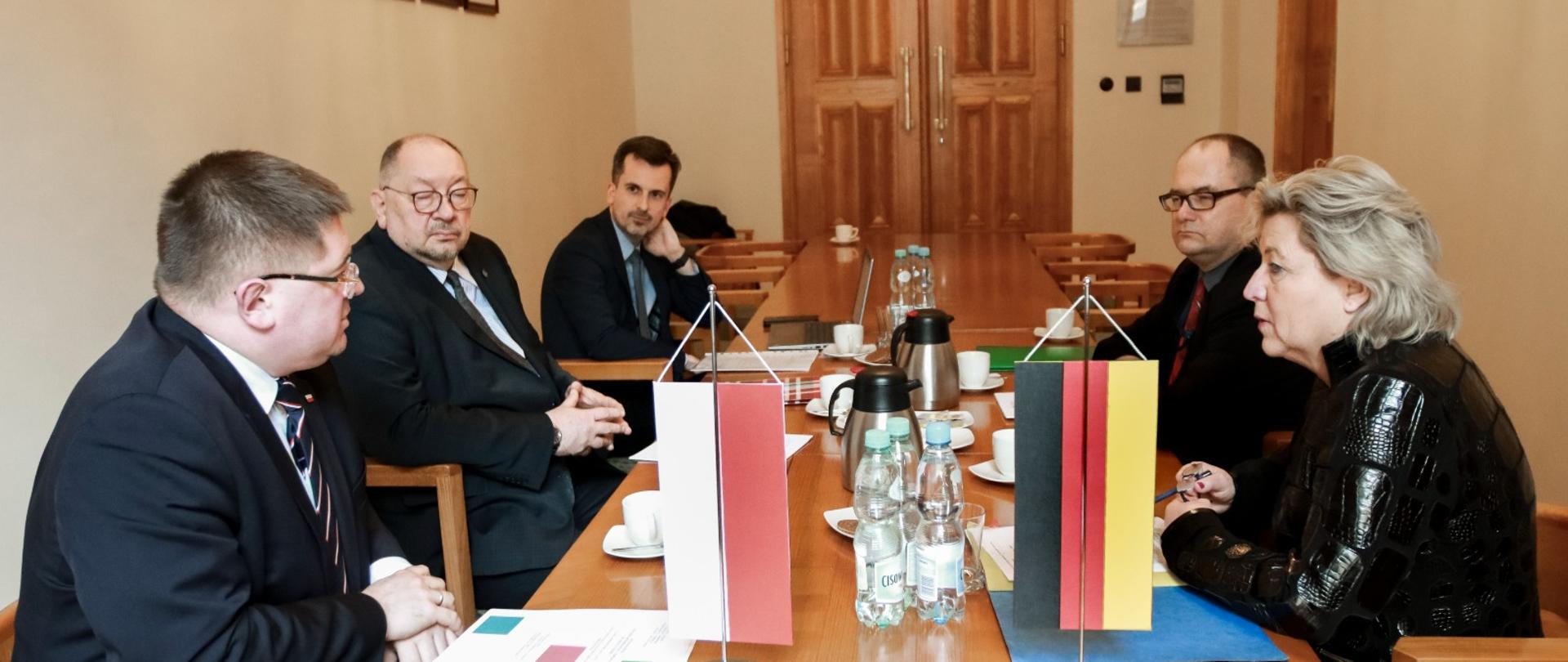 Widok z boku na czworokątny drewniany stół, przy którym siedzi pięć osób i rozmawia, za nimi białe ściany na których wiszą rzędy portretów, na środku stołu dwie małe flagi - polska i niemiecka, w głębi za stołem drewniane drzwi.