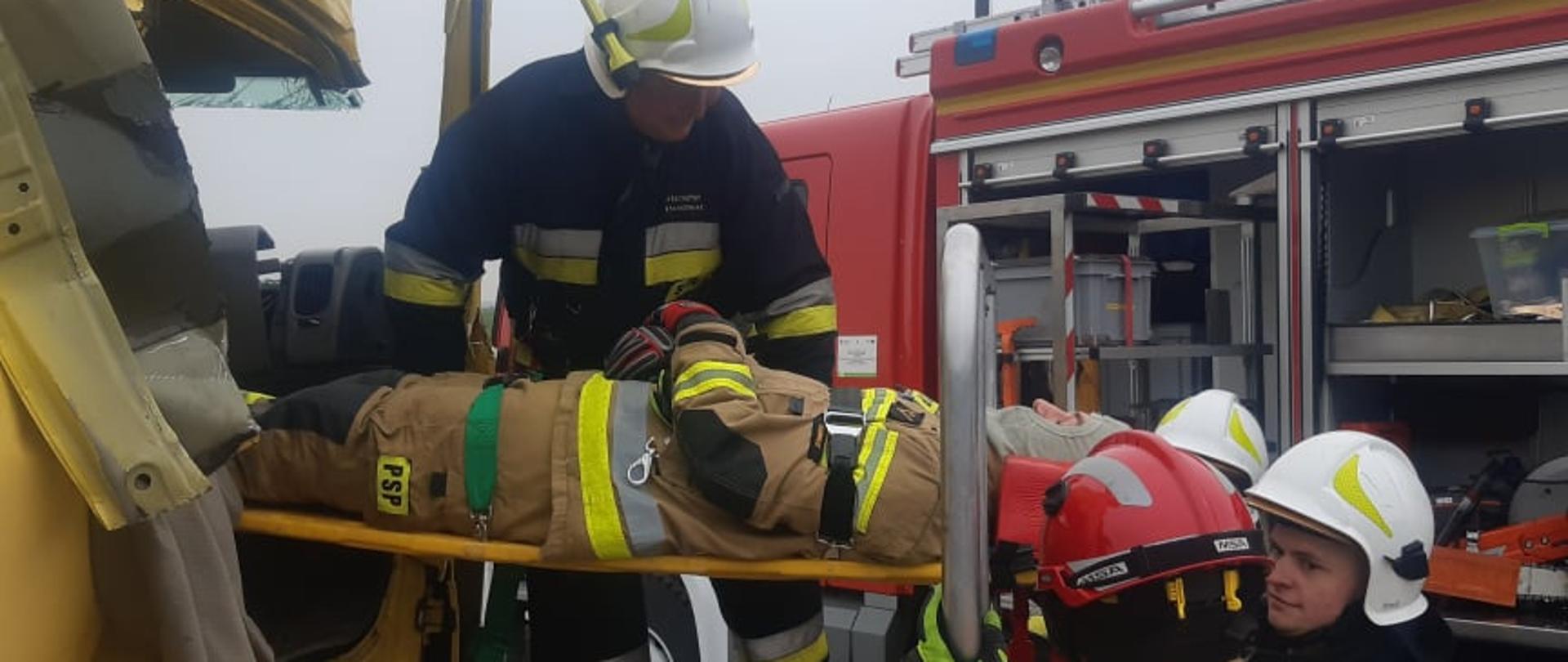 Warsztaty z zakresu ratownictwa technicznego. Złomowisko w Chojnie. Strażacy, którzy wcześniej wykonali dostęp do poszkodowanego znajdującego się w kabinie ciężarówki, ewakuują go na noszach typu deska. Jeden ze strażaków stoi na podeście ratowniczym, trzej pozostali na ziemi. W tle wóz straży pożarnej.