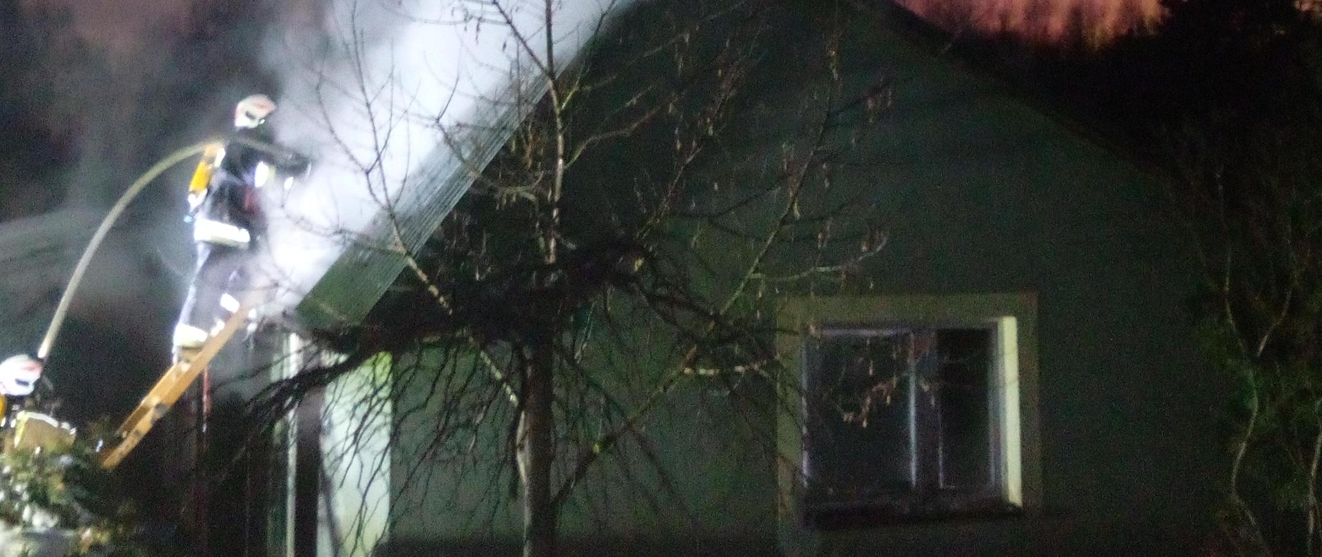 Zdjęcie przedstawia budynek gospodarczy, w którym doszło do pożaru. Na drabinie przystawionej do dachu stoi strażak podający strumień wody do wnętrza obiektu, a nad budynkiem unoszą się kłęby dymu i pary wodnej.