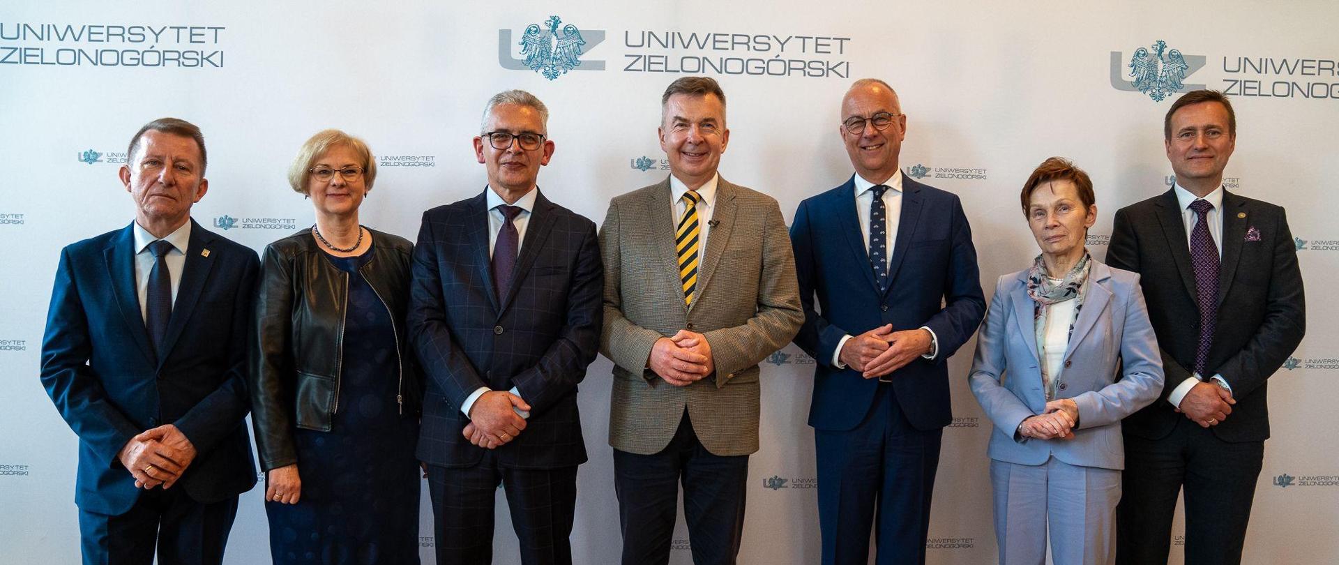 Minister Dariusz Wieczorek stoi w otoczeniu kobiet i mężczyzn elegancko ubranych. W tle napis Uniwersytet Zielonogórski.