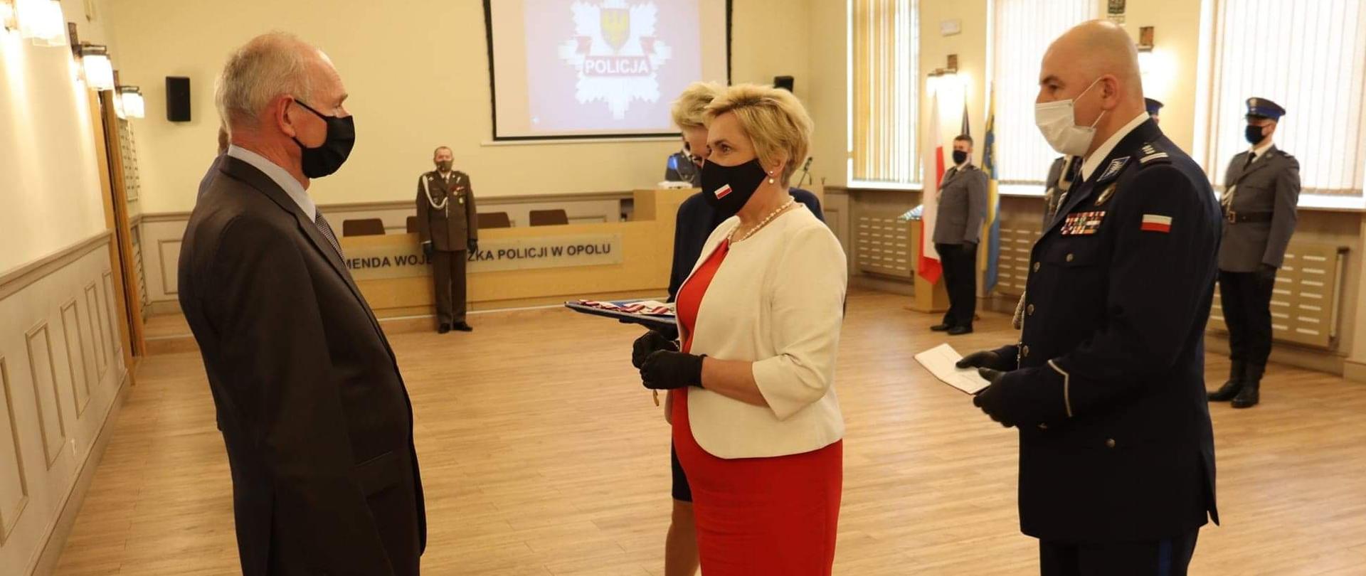 Wicewojewoda opolski ubrana w czerwoną sukienkę stoi na środku kadru w sali odznaczeń w opolskiej policji. Na przeciw wicewojewody stoi mężczyzna. W dłoniach wicewojewody odznaczenie państwowe. Obok wicewojewody po prawej stronie stoi policjant w mundurze. W tle pozostali uczestnicy uroczystości.