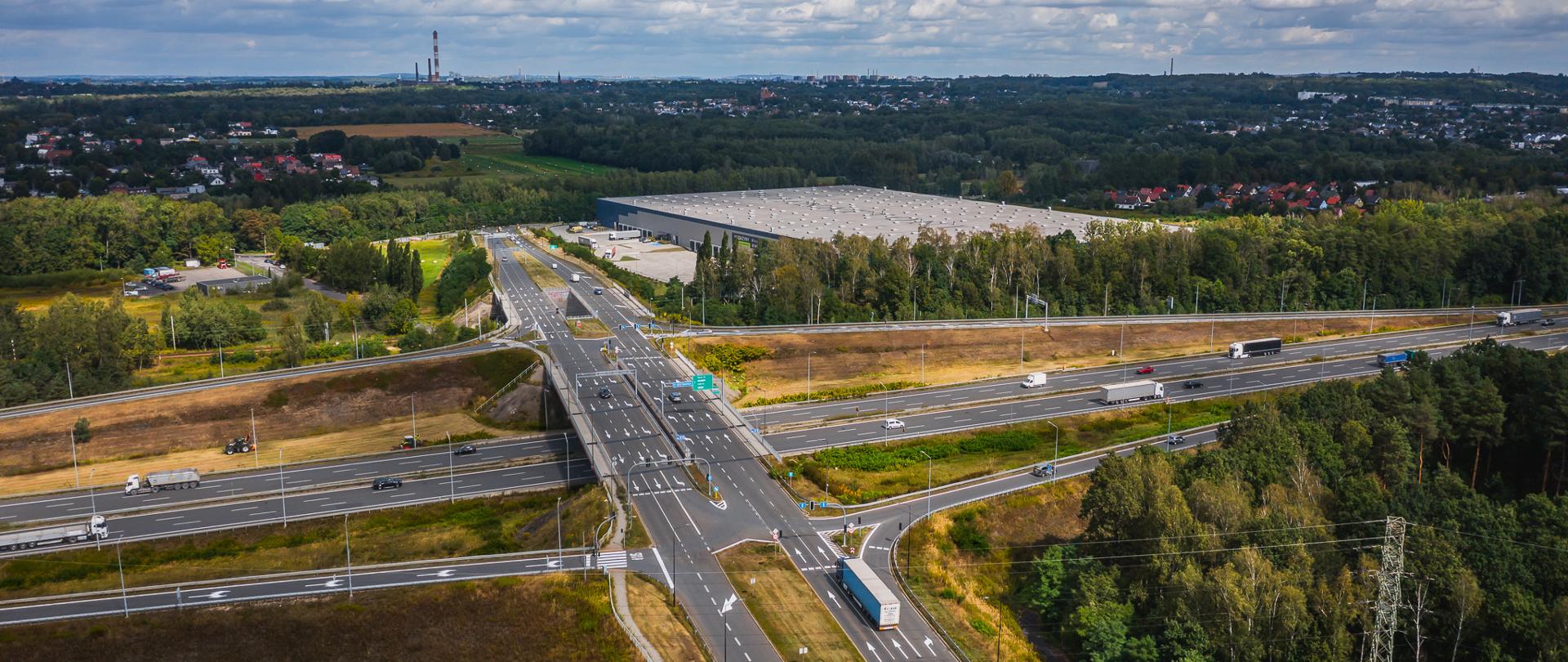 Panorama z lotu ptaka na układ drogowy, linię kolejową oraz hale magazynowe. Okoliczny teren porośnięty jest lasem oraz krzewami.