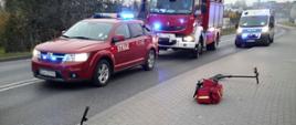 Na chodniku leży przewrócony rower, za nim torba ratownicza, dwa hełmy strażackie oraz przewrócona hulajnoga. W tle widać dwa samochody strażackie w włączonymi sygnałami świetlnymi oraz jeden policyjny.
