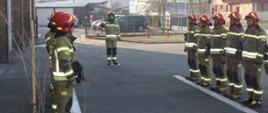 Zdjęcie przedstawia strażaków w umundurowaniu specjalnym stojących w dwóch szeregach naprzeciwko siebie. Jeden ze strażaków stojący w odległości ok. 2 m metrów od tych szeregów, salutuje. 