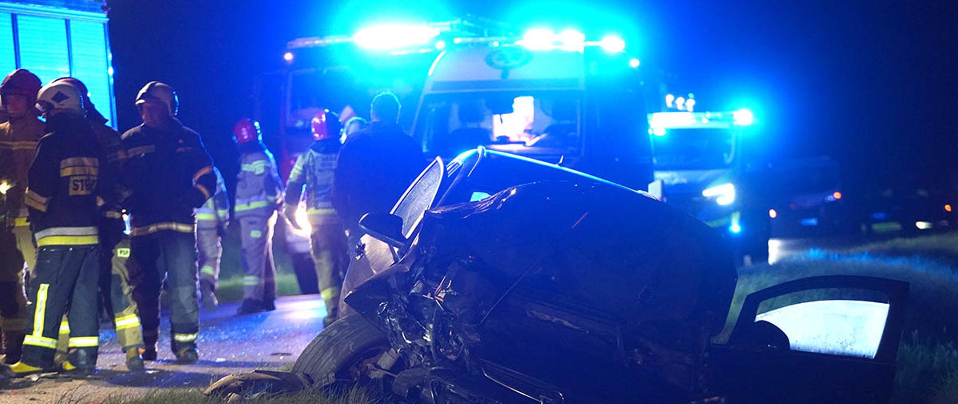 Rozbity samochód osobowy stoi na poboczu drogi nocą. Przód pojazdu jest poważnie uszkodzony. W tle widać strażaków i ratowników pracujących przy miejscu zdarzenia oraz pojazdy służb ratunkowych z włączonymi niebieskimi światłami.