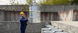 Pracownik ZUOP stojący w betonowym sektorze ustawia stalowy bęben z odpadami promieniotwórczymi. Bęben opuszczany jest przez dźwig.