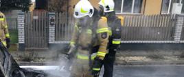 Zdjęcie na drodze publicznej. Widać auto, które uległo spaleniu. Wokół auta umundurowani ratownicy. Na drodze pozostałości piany gaśniczej. Zdjęcie wykonane w ciągu dnia.