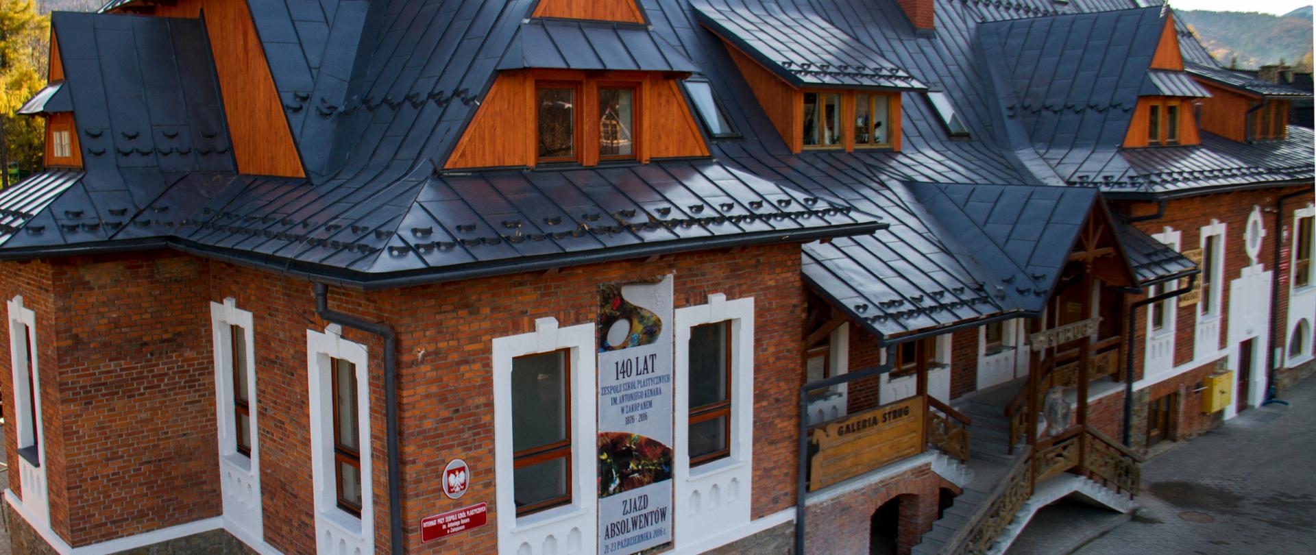 Zdjęcie przedstawia budynek Państwowego Liceum Sztuk Plastycznych im. Antoniego Kenara w Zakopanem. Elewacja wykonana jest z cegły, obramowania okien i dekoracja pod oknami wykonana jest na biało z tynku. Budynek posiada trzy kondygnacje. Poddasze jest drewniane. Całość pokryta jest spadzistym dachem, pokrycie dachu stanowi szara blacha. Internat dla 28 osób wydzielony jest z części budynku szkoły i na zdjęciu znajduje się po lewej stronie.