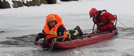 widać dwóch strażaków ubranych w kamizelki ratunkowe, jeden z nich leży na saniach lodowych, drugi który je pcha też jest w wodzie, bo załamał się pod nim lód.