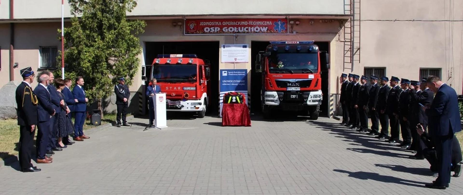 Plac przed remizą OSP w centrum dwa wozy strażackie stojące przodem, między nimi stojąca na stoliku torba PSP R-1, po bokach w dwóch rzędach stoją uczestnicy uroczystości - strażacy i zaproszeni goście