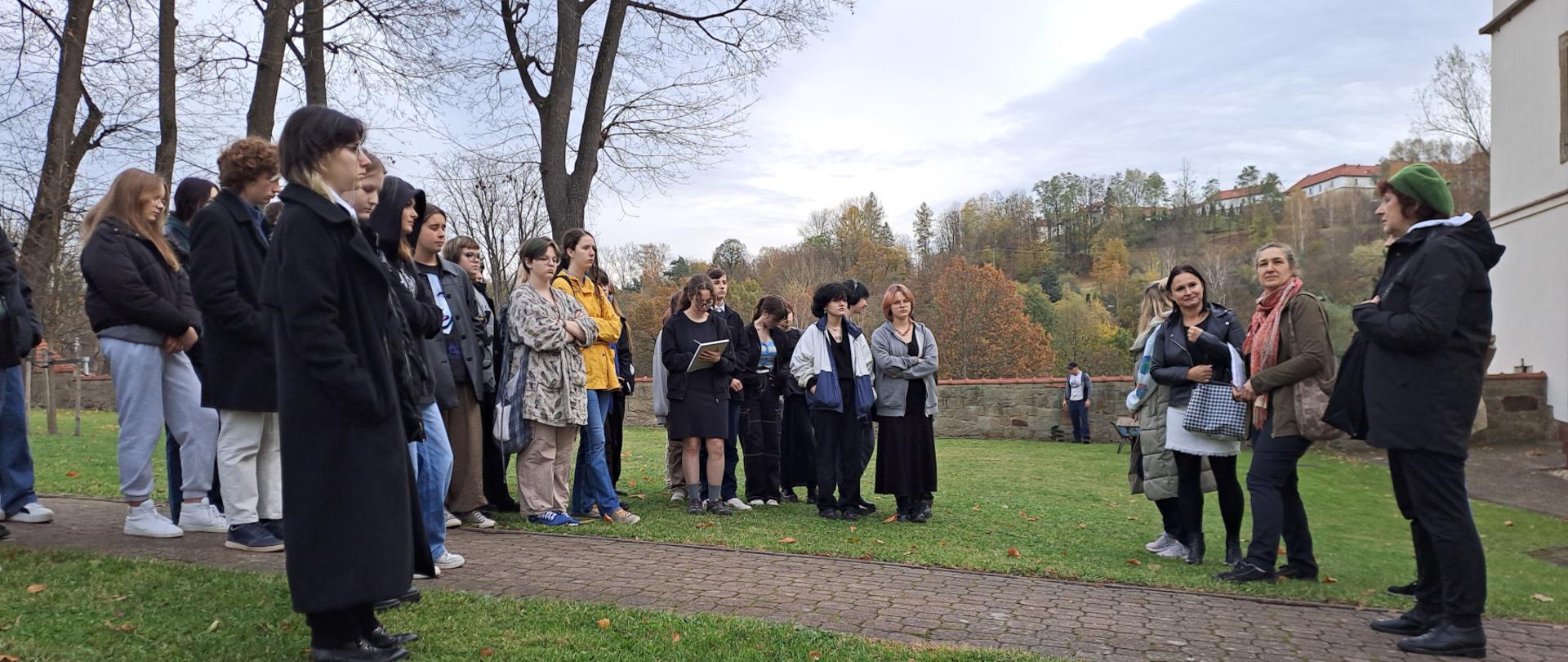 grupa młodzieży szkolnej stoi na trawniku koło kamiennej dzwonnicy i muru kościelnego zwrócona w stronę mówiącej pani stojącej po prawej stronie na pierwszym planie 