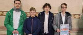 Czterech uczniów, w kurtkach i galowych koszulach stoi na tle stylizowanej ściany. Pierwszy z lewej trzyma w dłoniach białą papierową torbę, pierwszy z prawej dyplom zdobyty podczas konkursu. Napi na dyplomie - Bronzove Pasmo.