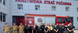 Grupa strażaków i uczestników kursu OSP stoi przed budynkiem Państwowej Straży Pożarnej. W tle widoczny jest czerwony wóz strażacki z napisem promującym bezpieczeństwo i czujniki tlenku węgla. Na elewacji budynku duży napis „PAŃSTWOWA STRAŻ POŻARNA”. Osoby na zdjęciu ubrane są w różne wersje mundurów – niektórzy w czarne bojowe, inni w piaskowe mundury nowego wzoru ubrań dla PSP i OSP. Grupa pozuje do wspólnego zdjęcia na brukowanym placu, wszyscy są uśmiechnięci, a pogoda jest słoneczna.