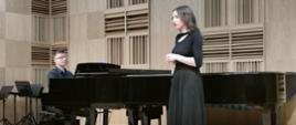Na scenie stoi młoda dziewczyna. Śpiewa do akompaniamentu fortepianu, na którym gra młody nauczyciel.