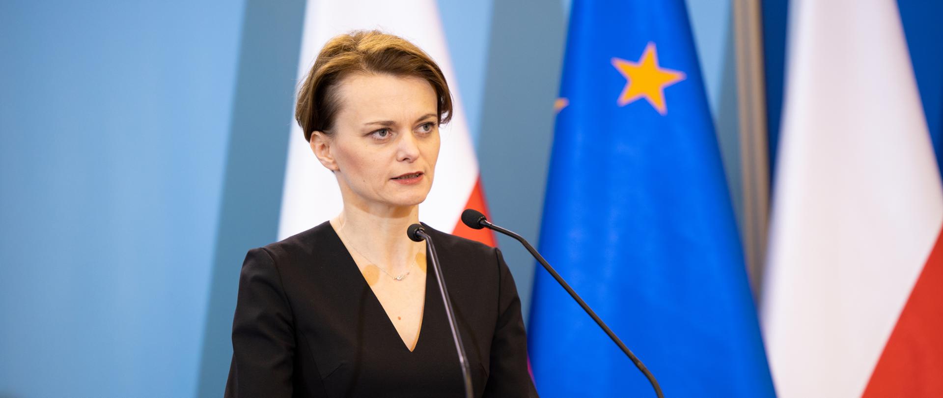 Wicepremier Jadwiga Emilewicz stoi za mównicą, za nią rozmyte flagi Polski i Unii Europejskiej.