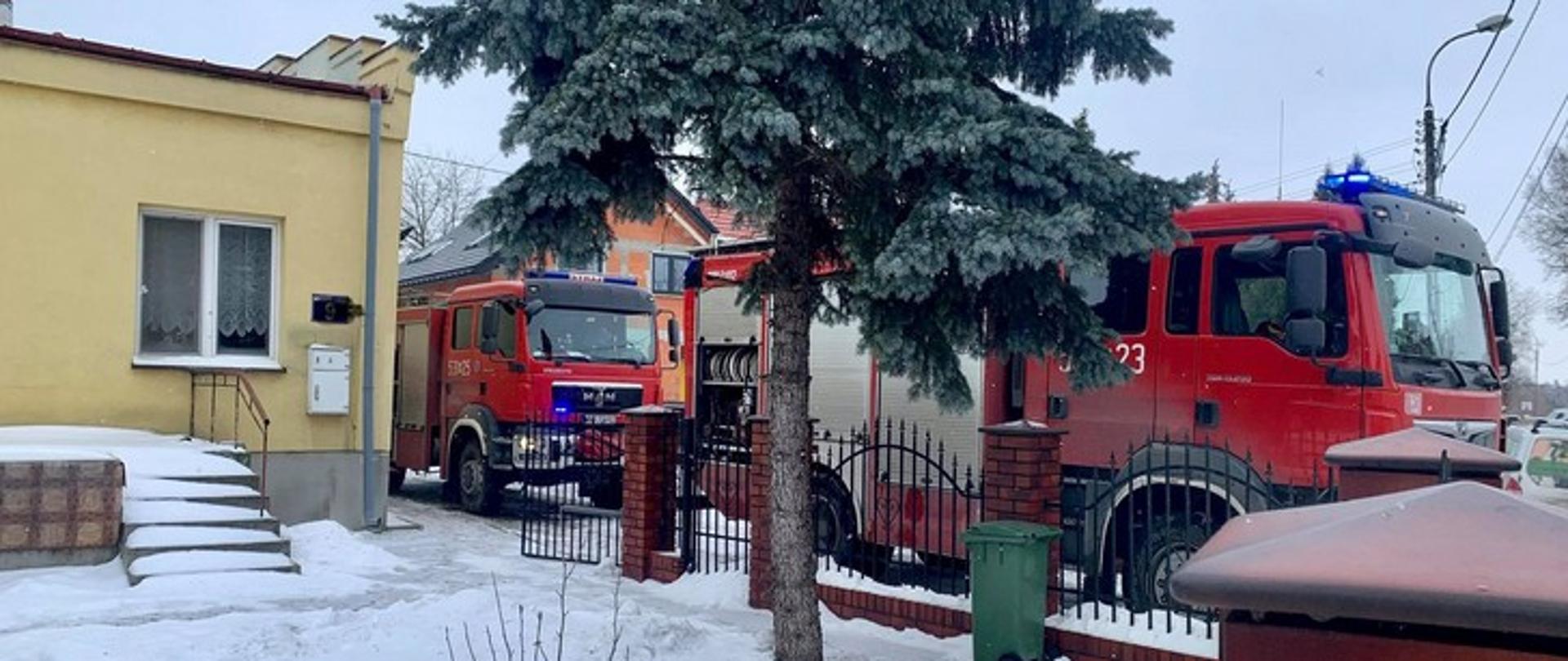 Zdjęcie przedstawia dwa samochody strażackie z włączonymi niebieskimi sygnałami ostrzegawczymi stojące wzdłuż ogrodzenia przy budynku mieszkalnym.