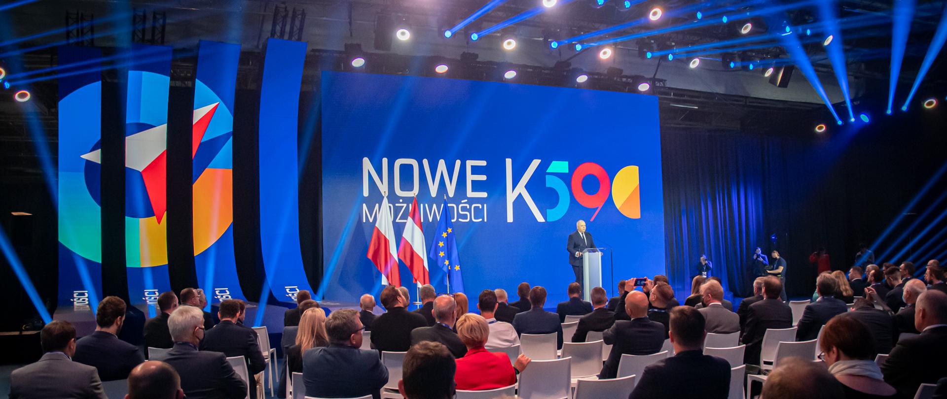 Szerokie ujęcie sali zza krzeseł z słuchaczami, w tle wicepremier przy mównicy, flagi, ścianka z napisem Nowe możliwości K590