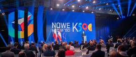 Szerokie ujęcie sali zza krzeseł z słuchaczami, w tle wicepremier przy mównicy, flagi, ścianka z napisem Nowe możliwości K590