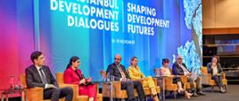 Grupa panelistów siedzi na fotelach, przemawia kobieta w rożowym garniturze, z tyłu widać napis Istanbul Development Dialogues 