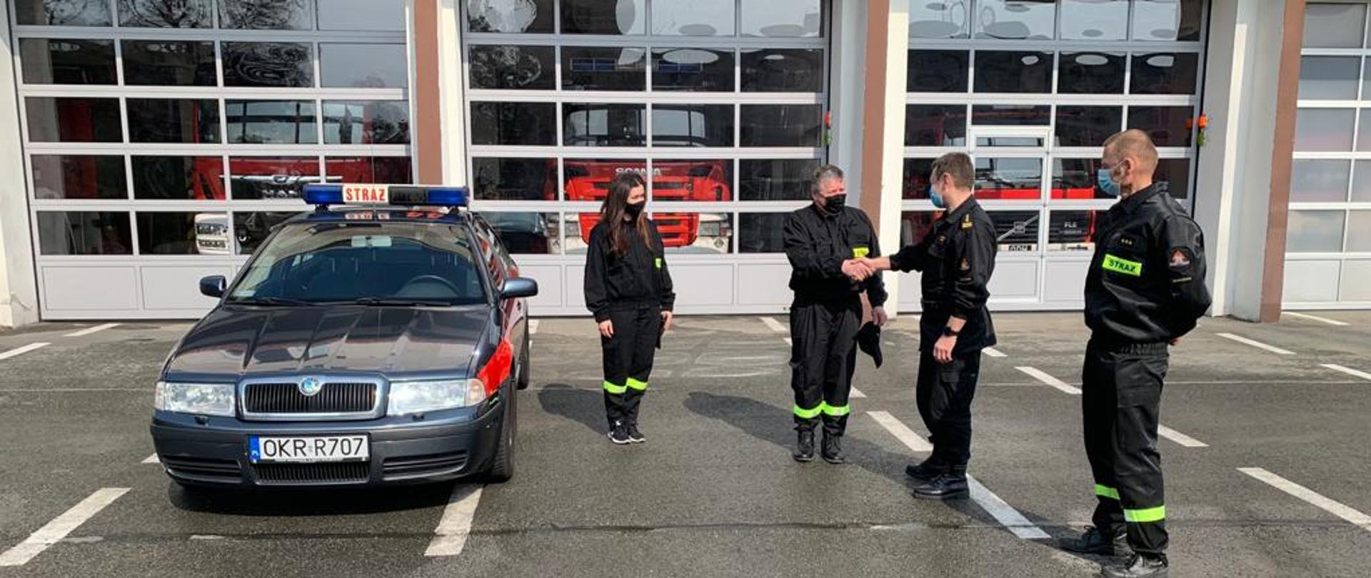 Na tle bram garażowych ustawiony jest przodem samochód operacyjny marki skoda octavia (lekko na lewo od osi zdjęcia). W środku i w kierunku w prawo ustawieni są: dwoje członków OSP - kobieta i mężczyzna, a następnie dwóch strażaków PSP. Strażacy OSP w ubraniach koszarowych i maseczkach. Pierwszy od lewej strażak PSP - Komendant Powiatowy w umundurowaniu dowódczo-sztabowym, drugi - kierowca w ubraniu koszarowym. Na twarzach mają założone maseczki. Komendant Powiatowy podając dłoń gratuluje prezesowi OSP otrzymanego samochodu. 