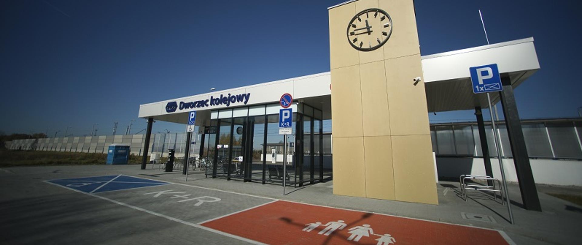 Na zdjęciu dworzec kolejowy, szklana wiata, wieża z zegarem, miejsca parkingowe dla osób z niepełnosprawnościami, rodzin z dziećmi oraz tzw. kiss and ride.