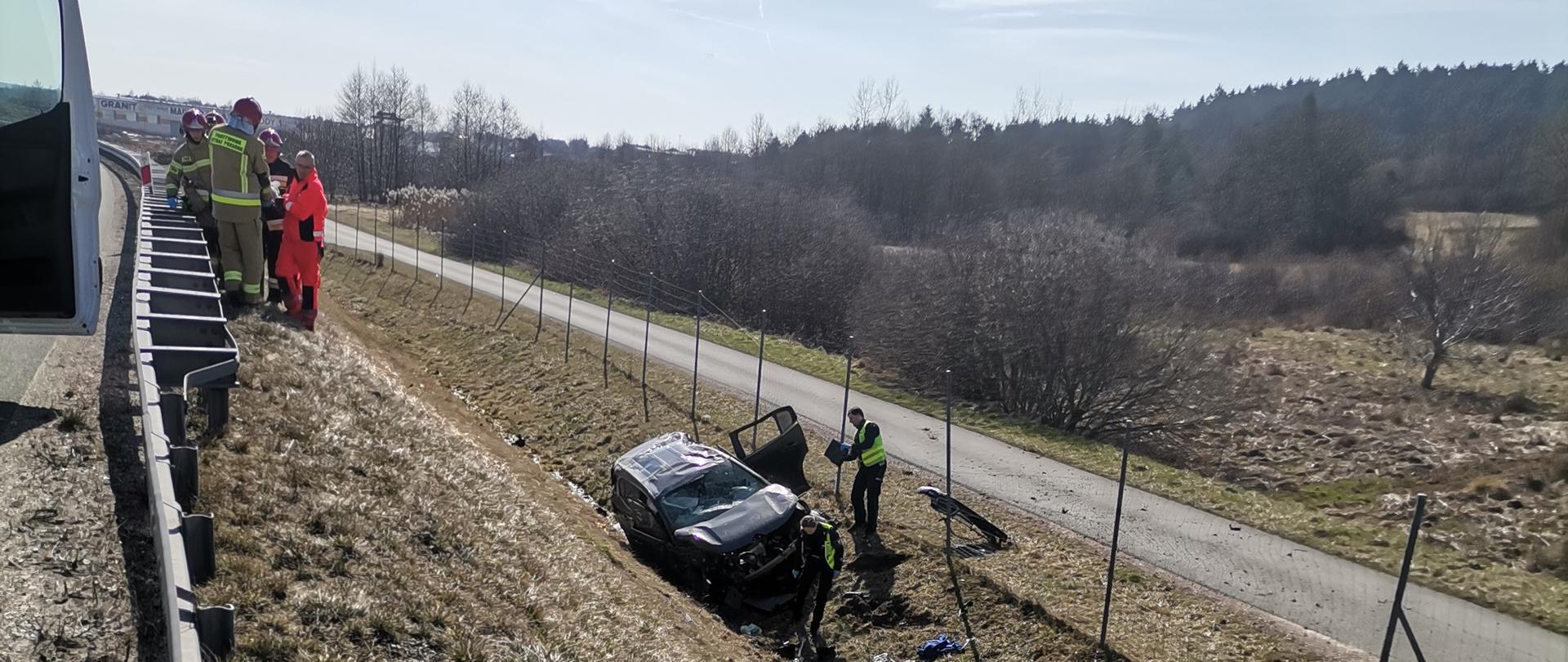 Zdjęcie przedstawia głęboki rów przydrożny, w którym znajduje się uszkodzony samochód osobowy. Przy nim funkcjonariusze Policji prowadzą działania. Po lewej stronie, na koronie nasypu widać stojących strażaków z ratownikami medycznymi.