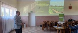 Sala szkoleniowa podczas prezentacji dotyczącej rolnictwa i ochrony roślin. Na pierwszym planie stoi mężczyzna w jasnoszarej marynarce i ciemnych spodniach, prowadzący spotkanie. Za nim, na ścianie, wyświetlony jest slajd prezentacji z fotografią pól uprawnych oraz tekstem dotyczącym stosowania środków ochrony roślin sprzętem naziemnym. W dolnej części zdjęcia widoczny jest stół organizatorów z otwartym laptopem, projektorem, dokumentami i jednorazowym kubkiem. W sali znajdują się drewniane stoły i krzesła, a uczestnicy siedzą po prawej stronie i słuchają wystąpienia. Wnętrze jest przestronne, jasne i utrzymane w prostym, użytkowym stylu.