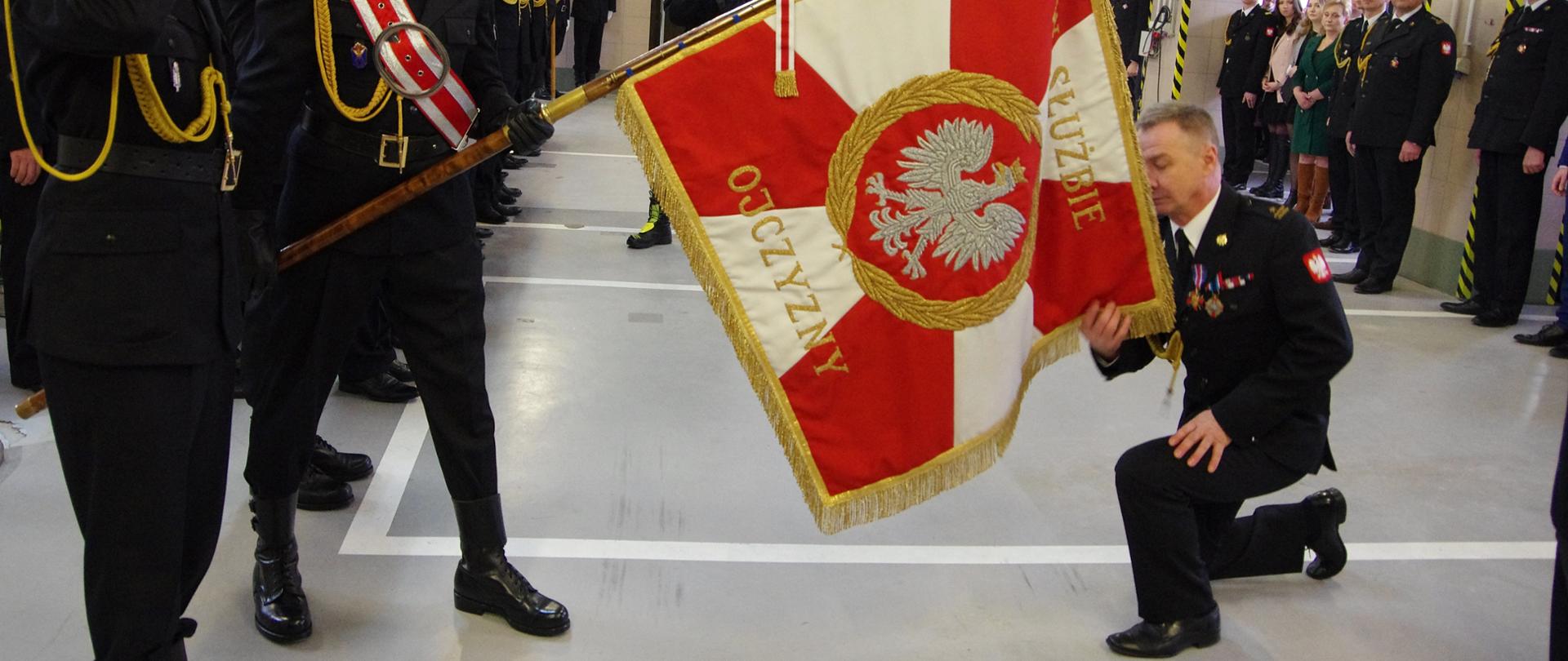 Strażak przyklęka na jedno kolano i całuje sztandar. W tle uczestnicy uroczystej zbiórki.