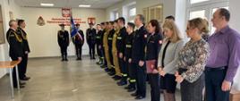 Na zdjęciu w mundurach galowych po lewej komendant powiatowy wraz z zastępcą po środku poczet sztandarowy ze sztandarem. Po prawej strażacy ubraniu czarne i piaskowe ubrania koszarowe oraz ubrania galowe a także pracownicy cywilni komendy uczestniczący w uroczystość ślubowania na sali konferencyjnej.