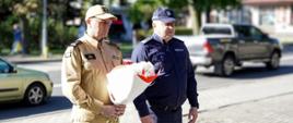 Zdjęcie przedstawia komendanta straży i komendanta policji z Ustrzyk Dolnych. Komendant straży pożarnej trzyma w rękach kwiaty. Obaj stoją na rynku w Ustrzykach Dolnych.