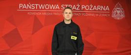 Strażak w czarnym mundurze stoi na tle czerwonej ścianki z napisem ‘Państwowa Straż Pożarna – Komenda Miejska Państwowej Straży Pożarnej w Żorach’ oraz logo PSP. Patrzy prosto w kamerę i trzyma dokument w prawej ręce
