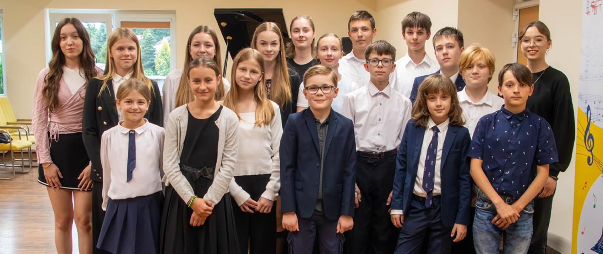 Na zdjęciu grupa kilkunastu osób, głównie dzieci stoi w auli szkoły, za nimi stoi fortepian.