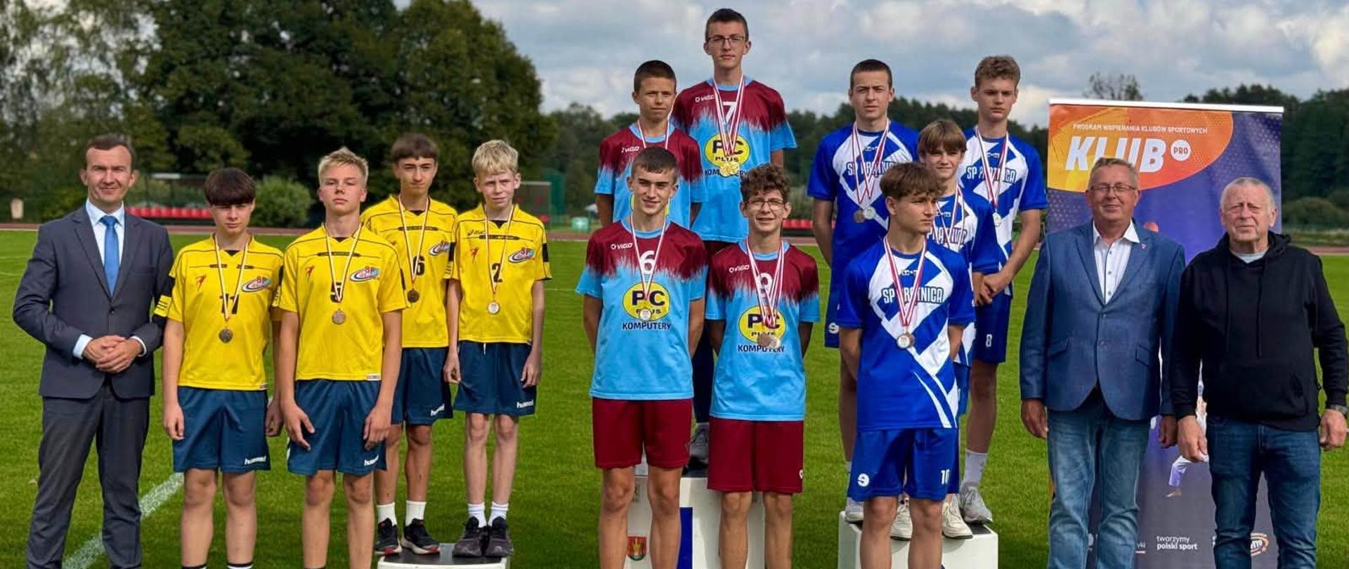 Trzy stopnie podium zajmują różne grupy sportowe w odmiennych koszulkach. Obok nagrodzonych zawodników stoi wicewojewoda w szarym garniturze.