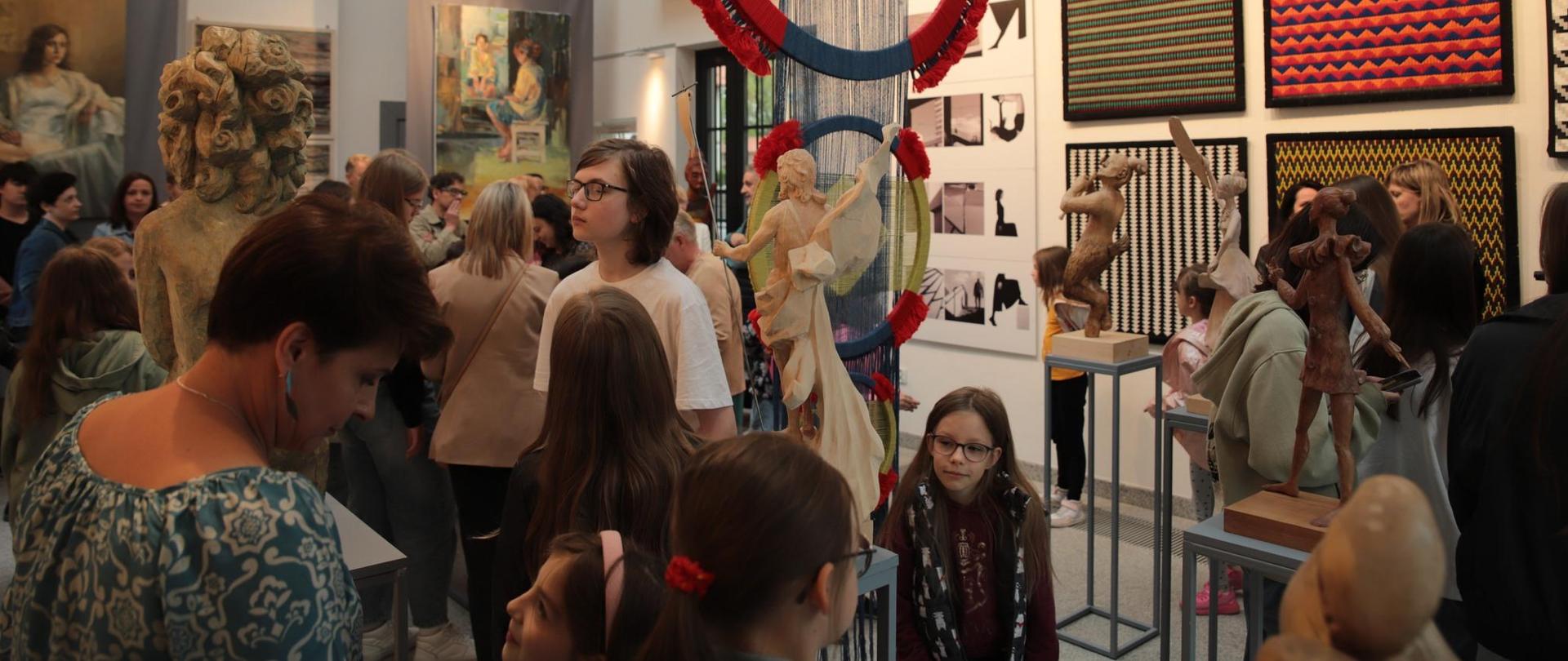 Sala wystawowa w Domu Kultury w Lubartowie. Jest wysoka. W suficie są skosy, tam znajdują się okna dachowe (weluxy). Przy suicie dwa szeregi świateł - reflektorów. W sali znajdują się prace plastyczne z różnych specjalizacji: malarstwo, tkanina artystyczna, rzeźba, projektowanie graficzne. Jest dużo zwiedzających w różnym wieku, począwszy od kilkuletnich dzieci, a skończywszy na osobach dorosłych, po 60-tce.