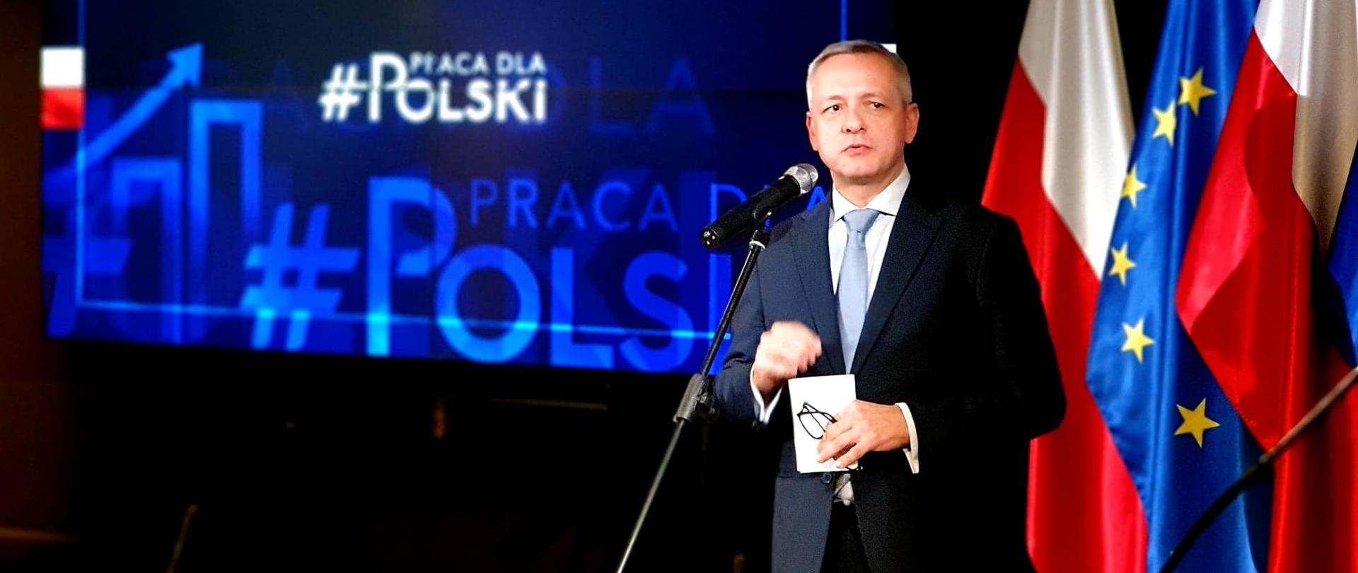 Minister cyfryzacji, Marek Zagórski, podczas konferencji prasowej przemawia do mikrofonu. W tle prezentacja multimedialna oraz flagi Polski i UE.