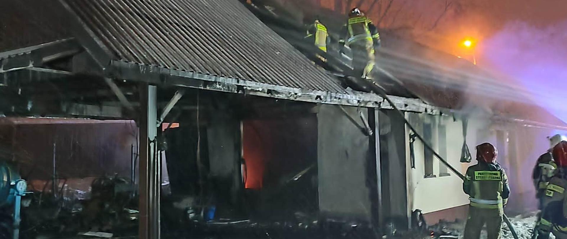 W nocy, przed budynkiem strażacy prowadzą działania gaśnicze podając wodę na dach. Na dachu w spalonej części stoją inni strażacy, którzy dogaszają wnętrze obiektu. 