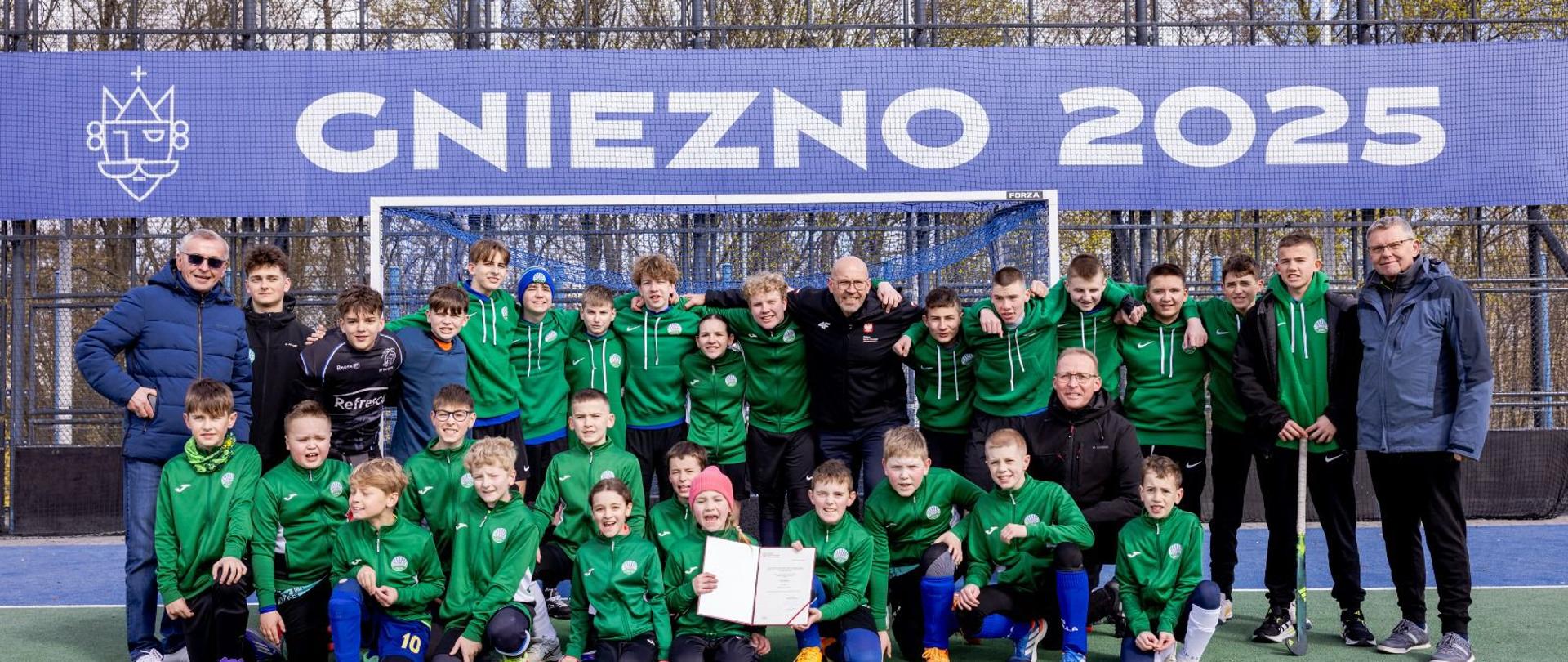 Minister Jakub Rutnicki na boisku do hokeja na trawie w Gnieźnie. Minister stoi z zawodnikami w zielonych bluzach na tle bramki i siatki