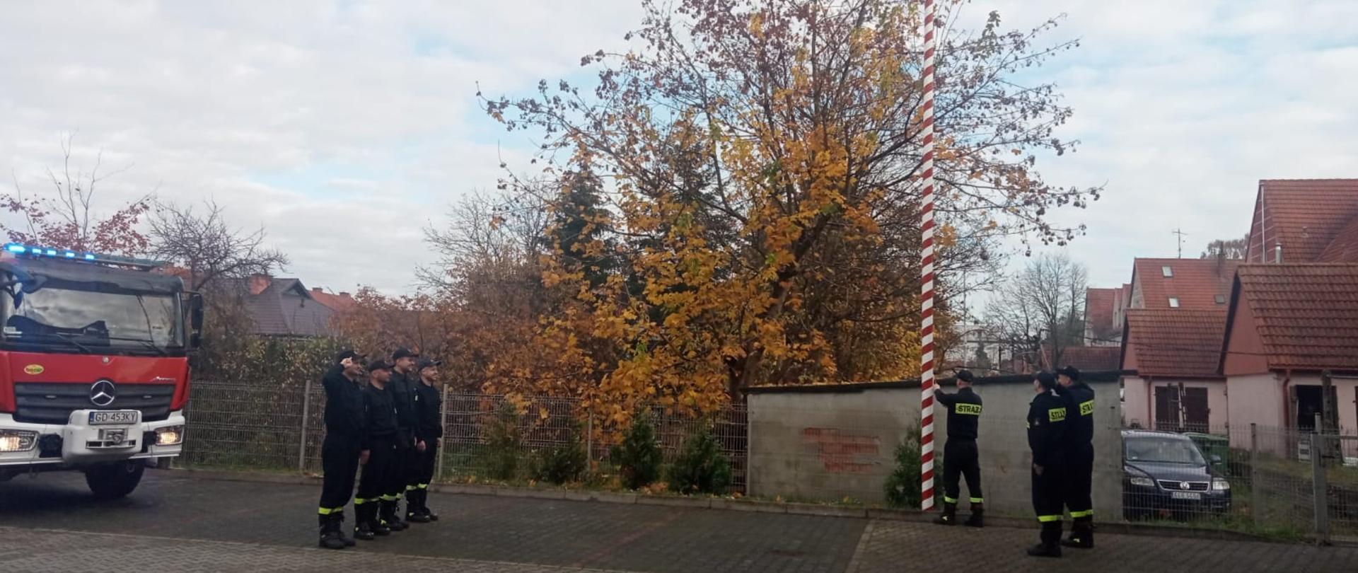 Strażacy uroczyście podnoszą flagę państwową na maszt. Sześciu strażaków stoi po dwóch stronach masztu na baczność. Trzech z nich salutuje. Jeden strażak wciąga flagę na maszt.