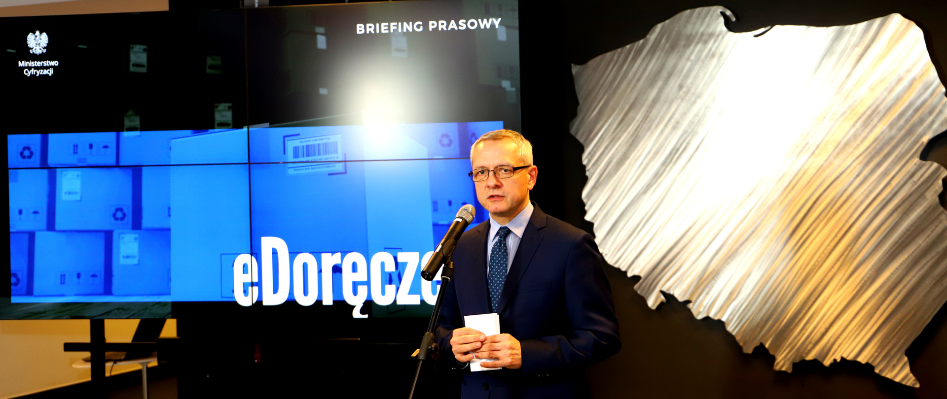 Minister Marek Zagórski podczas briefingu prasowego , za nim kontur mapy Polski oraz napis "eDoręczenia".