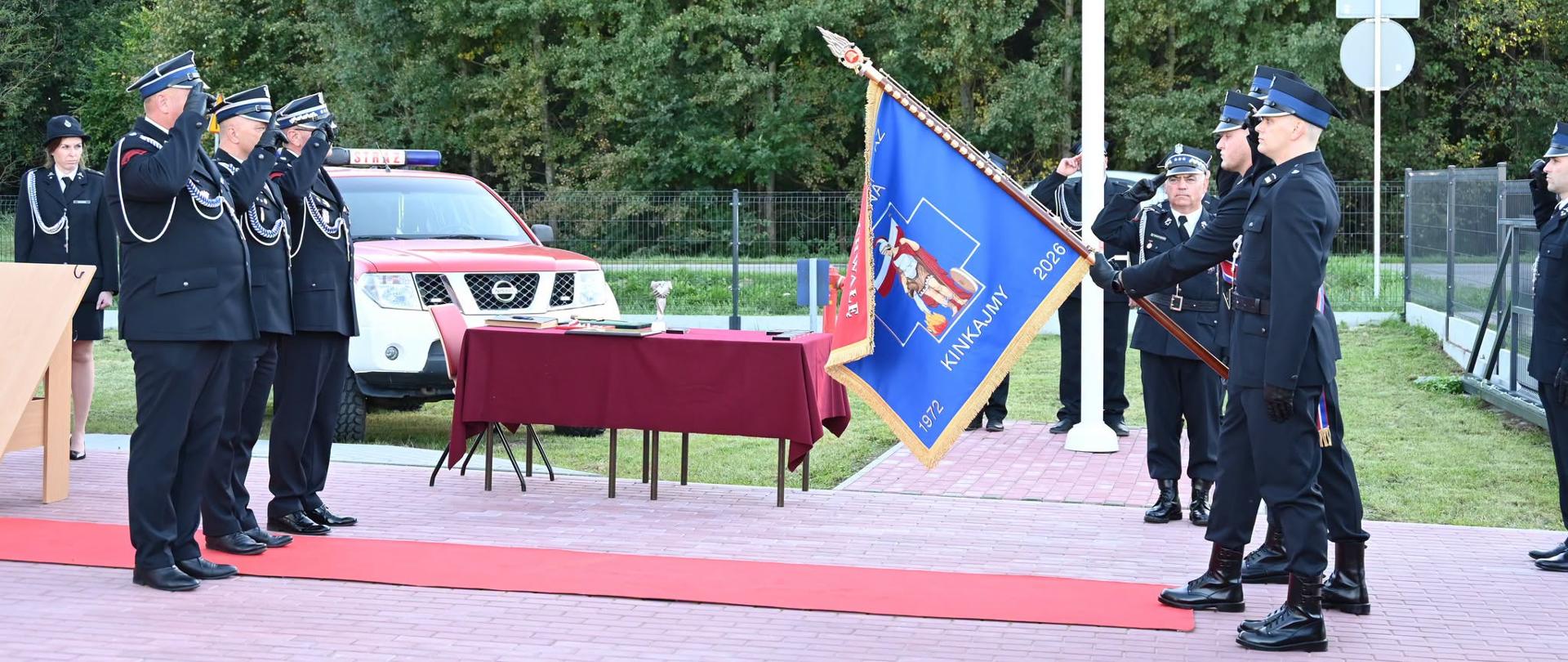 Oficjalne oddanie honorów sztandarowi. Poczet trzymający niebieski sztandar OSP Kinkajmy prezentuje go przed strażakami w mundurach galowych, którzy salutują. W tle widać czerwony stół i wóz strażacki.