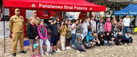 Zdjęcie strażaków z dziećmi podczas eventu promującego bezpieczeństwo na plaży. Czerwony namiot z napisem Państwowa Straż Pożarna. W tle zabudowania deptaku w kształcie parasoli.