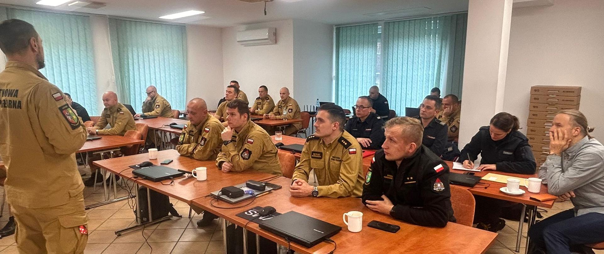 Instruktor stoi na środku sali i przemawia do uczestników. W ławkach siedzą polscy i niemieccy strażacy, a w rogu sali widać ułożone w stos kartony ze sprzętem komputerowym.