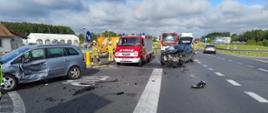 Na drodze widoczne są dwa uszkodzone samochody osobowe po zderzeniu. W pobliżu pojazdów pracują strażacy oraz znajdują się samochody służb ratowniczych. Jezdnia jest zabezpieczona, a na jej powierzchni widoczne są elementy uszkodzonych pojazdów.